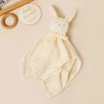 A soft bunny-shaped muslin security blanket in cream, styled on a warm beige background with a wooden ring toy and a round wooden sign in the background.