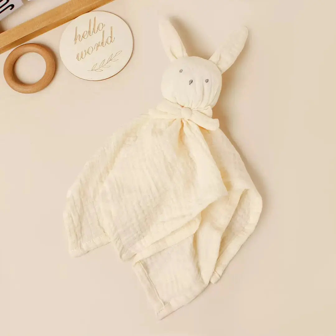 A soft bunny-shaped muslin security blanket in cream, styled on a warm beige background with a wooden ring toy and a round wooden sign in the background.