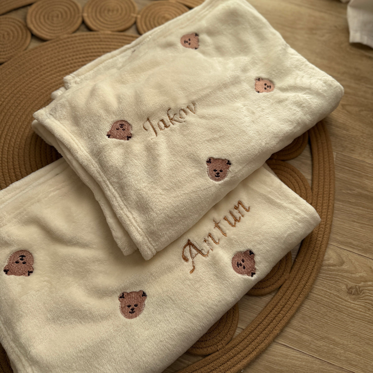 Two beige towels with embroidered teddy bear faces and names on a wooden floor.