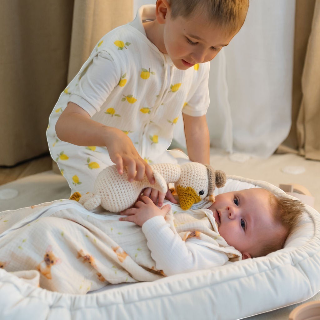 Baby Natur white sleeveless sleeping bag with yellow lemon print, worn by a boy. A baby lies in a bassinet with a knitted duck toy.