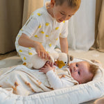 Baby Natur white sleeveless sleeping bag with yellow lemon print, worn by a boy. A baby lies in a bassinet with a knitted duck toy.