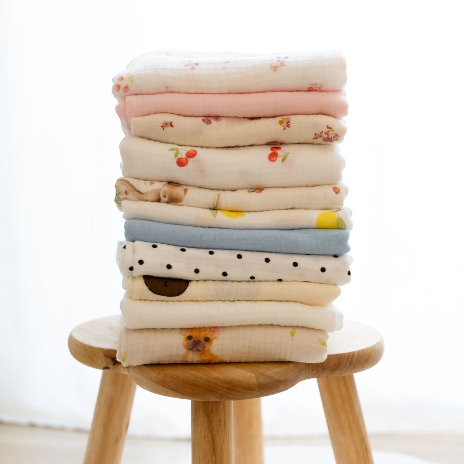 Stack of Baby Natur 100% cotton muslin cloths with various cute prints like cherries, bears, dogs, and polka dots.