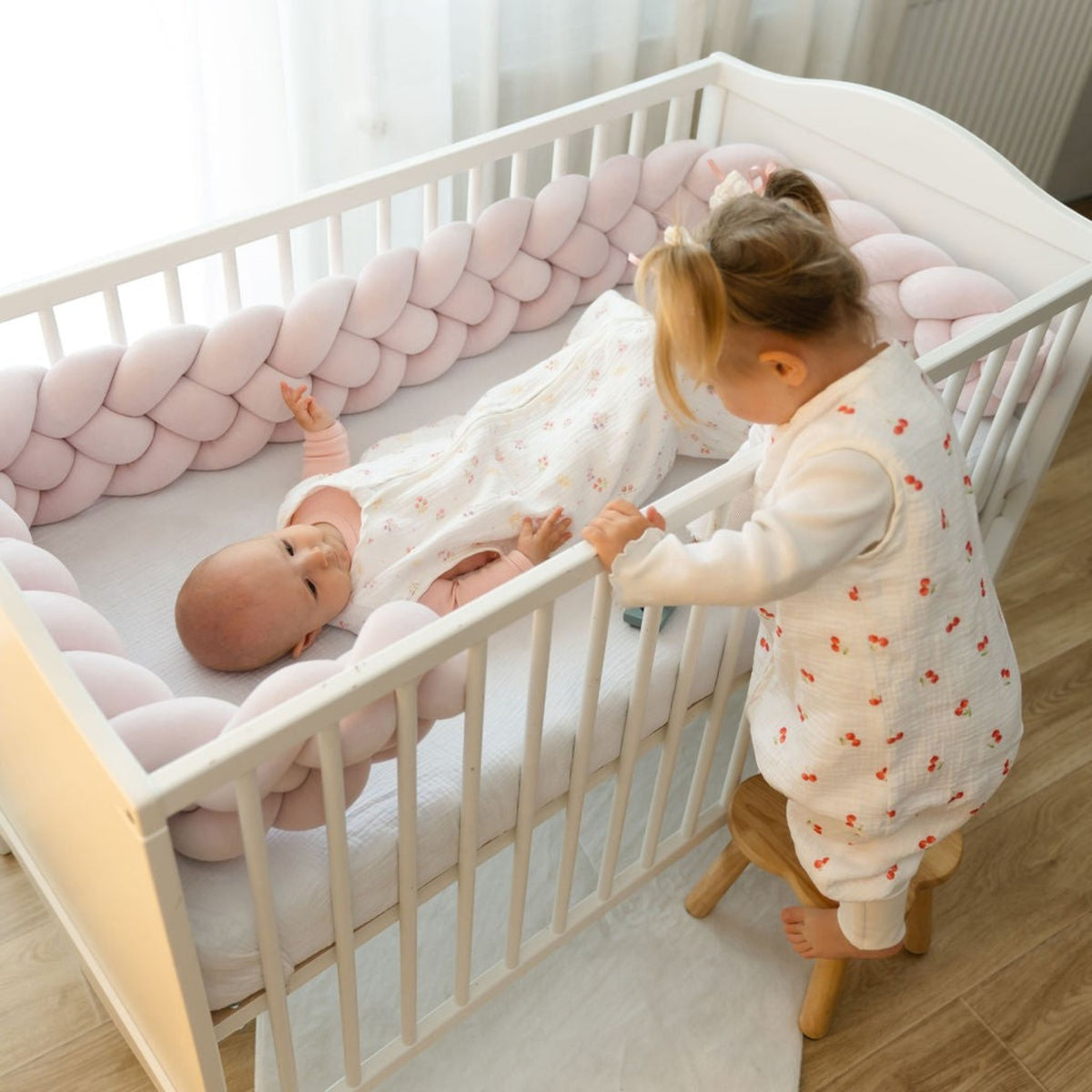 Baby Natur pink braided crib bumper with a baby sleeping inside and a toddler looking in.