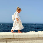 Baby Natur white muslin poncho with red cherry print, worn by a young girl walking by the sea.