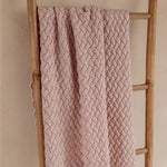 A dusty pink cable-knit blanket draped over a rustic wooden ladder against a neutral beige wall.